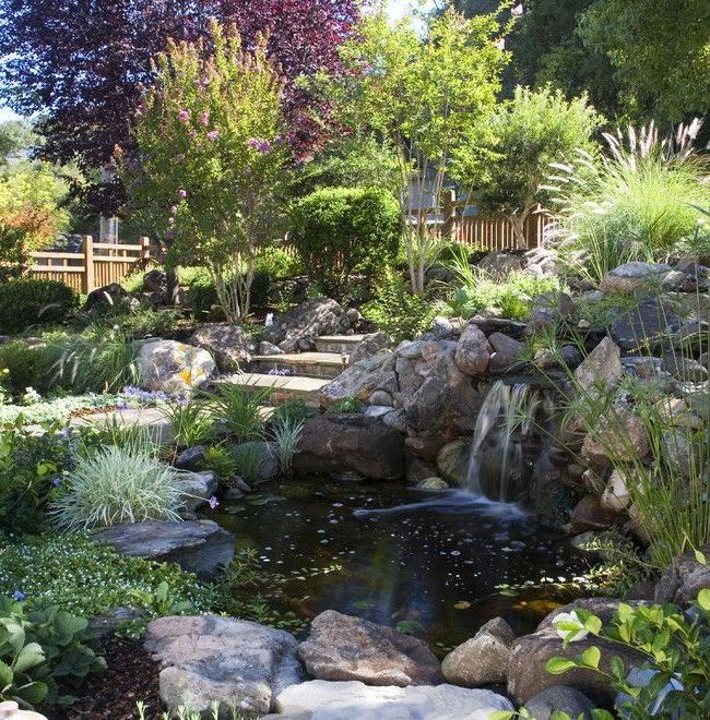 A pond surrounded by stones with a waterfall in a green garden